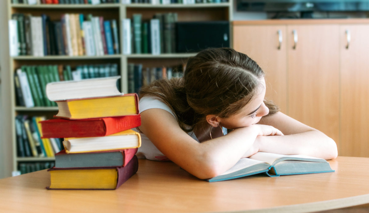 Student is asleep at a desk with their head on their hands, resting on an open book. There's a big pile of books next to her on the desk.