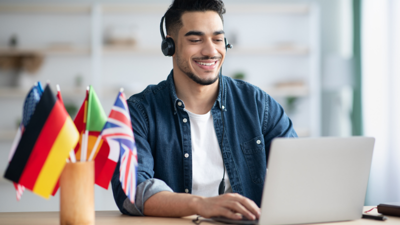 A student wearing headphones, typing on a laptop, learning a foreign language independently. On the desk are some country flags.