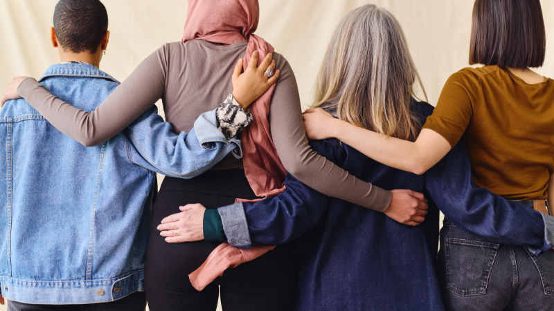 Rear portrait of four women standing in a line with their arms around each other.