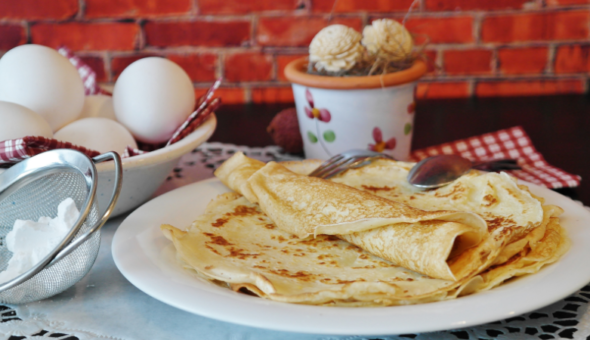 French crêpes on a plate next to a bowl of eggs and a sieve with flour in.