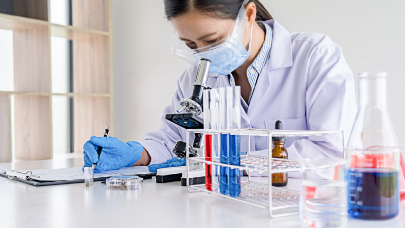 Person in a lab coat looking into a microscope doing an experiment in a laboratory. There's a row of test tubes on the bench. The person is writing on a clipboard.
