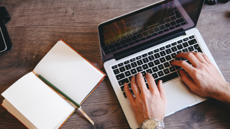 Hands resting on the keyboard of a laptop, with an open notebook and pencil to the left