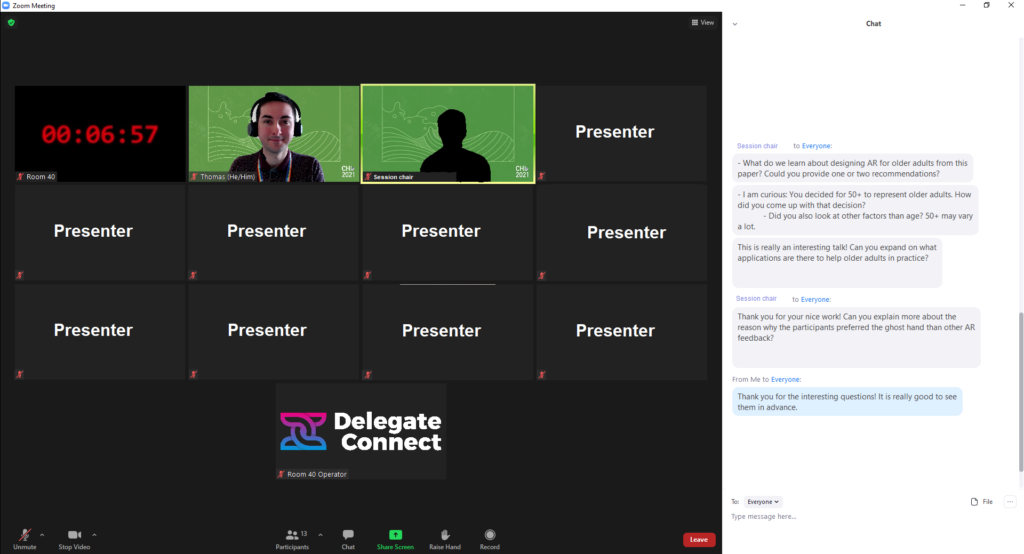 A screenshot of the presenter's experience. A Zoom meeting with 13 participants, shown in gallery view. I have my video on, and the chair of the session's video is displayed but with the presenter blacked out for anonymity. The top left participant is a count down, controlled by the service providing the technical support, Delegate Connect, whose logo is visible as another participant. All other presenters (9 in total) are anonymous authors with their cameras off. The chat is open and a sample of questions that I was asked by the session chair are visible. The questions are: 1. What do we learn about designing AR for older adults from this paper? Could you provide one or two recommendations? 2. I am curious: You decided for 50+ to represent older adults. How did you come up with that decision? 3. Did you also look at other factors other than age? 50+ may vary a lot. 4. This is really an interesting talk! Can you expand on what applications are there to help older adults in practice? 5. Thank you for your nice work! Can you explain more about the reason why the participants preferred the ghosthand than other AR feedback? A final comment from me reads: Thank you for the interesting questions! It is really good to see them in advance.