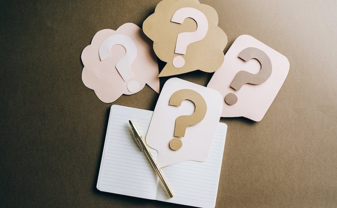 A white piece of paper and a pen on a grey table with paper question marks surrounding it.