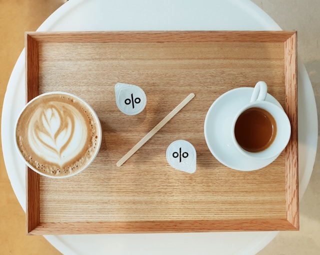 Two cups of coffee on a tray, with a stirring stick and two sachets of milk arranged to look like a percentage symbol.