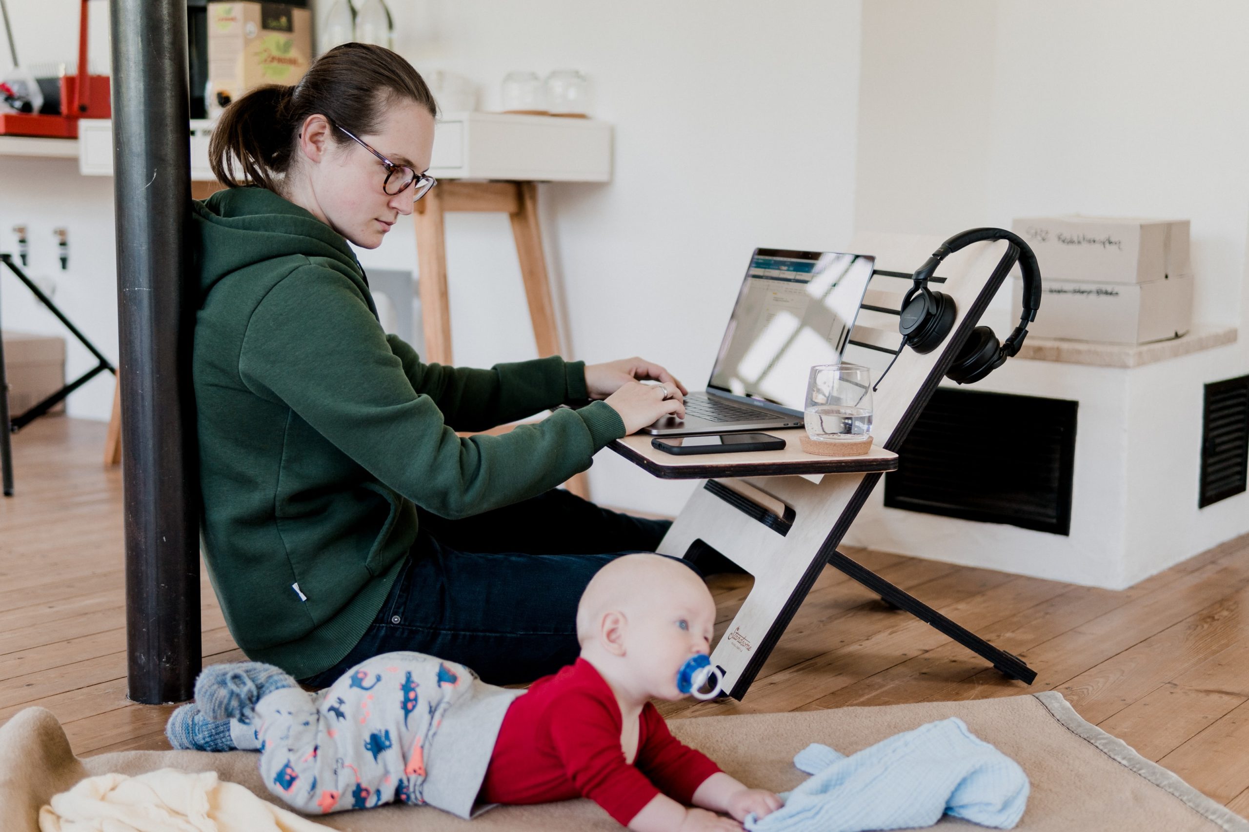 Woman with green hoody and spectacles sits on the floor at a modern desk with laptop. Her baby is to her right, has a dummy in its mouth and a bright red top.