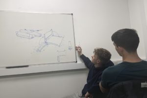 A white man sketching a military tank on a whiteboard, while another man is observing.
