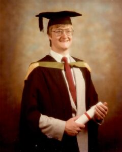 official graduation photo of david glass. Man in graduation gown and mortarboard smiling to camera with graduation certificate in hand