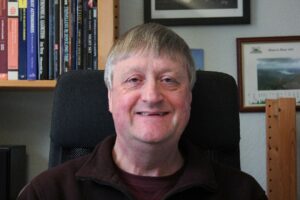 David Glass now. Man smiles to webcam on a computer chair with bookcases lining the walls behind him