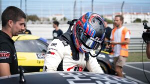 Filip on the race track stood next to a car with a helmet on.