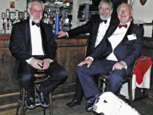 three men in suits and dicky bows sit on bar stools a bar in pub with a dog at their feet