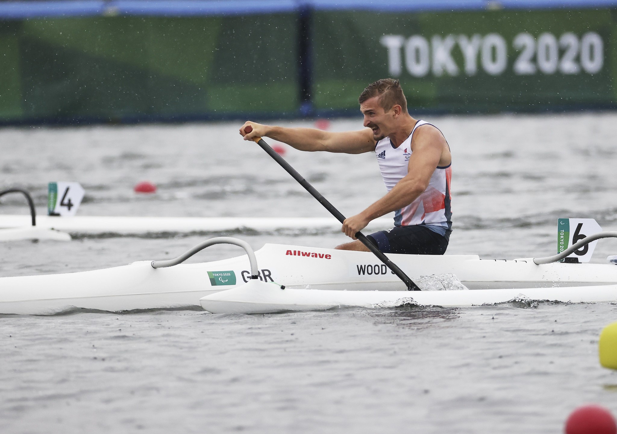 A male Paralympian rowing a canoe in a river, with a sign saying Tokyo 2020 in the background.