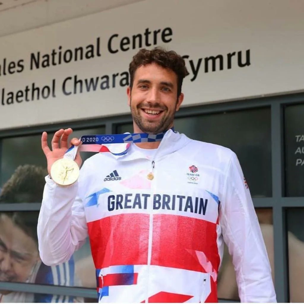 A brunette male with a beard wearing a white sports kit holding a gold Olympic medal.
