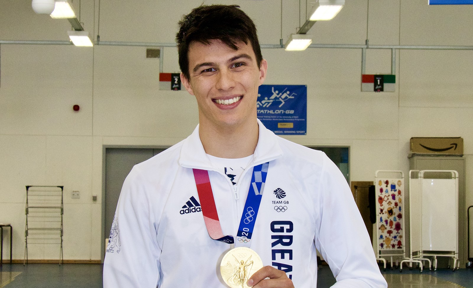 Brunette male in a white sports kit grinning and holding a gold Olympic medal.