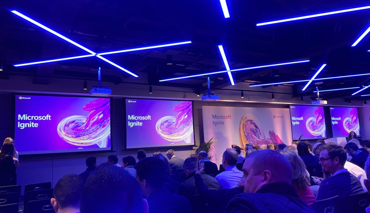 Conference venue with people seated in rows and large screens in the background with a Microsoft Ignite title and graphic.