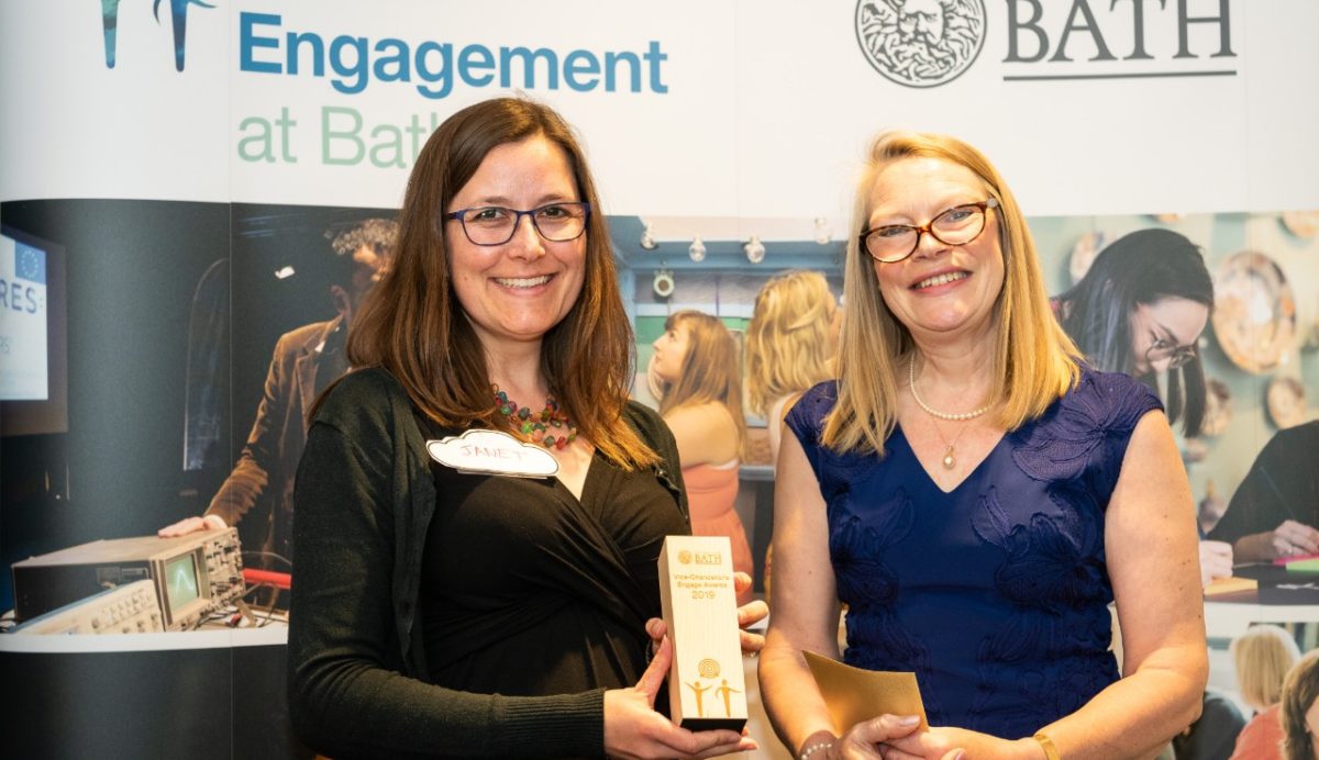 Two Caucasian women smiling at the camera. One is holding a small wooden trophy and they are standing in front of a large banner which reads 'Public Engagement at Bath'.