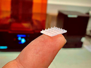 3D printed microneedles rest on a person’s fingertip in front of a 3D printer. 