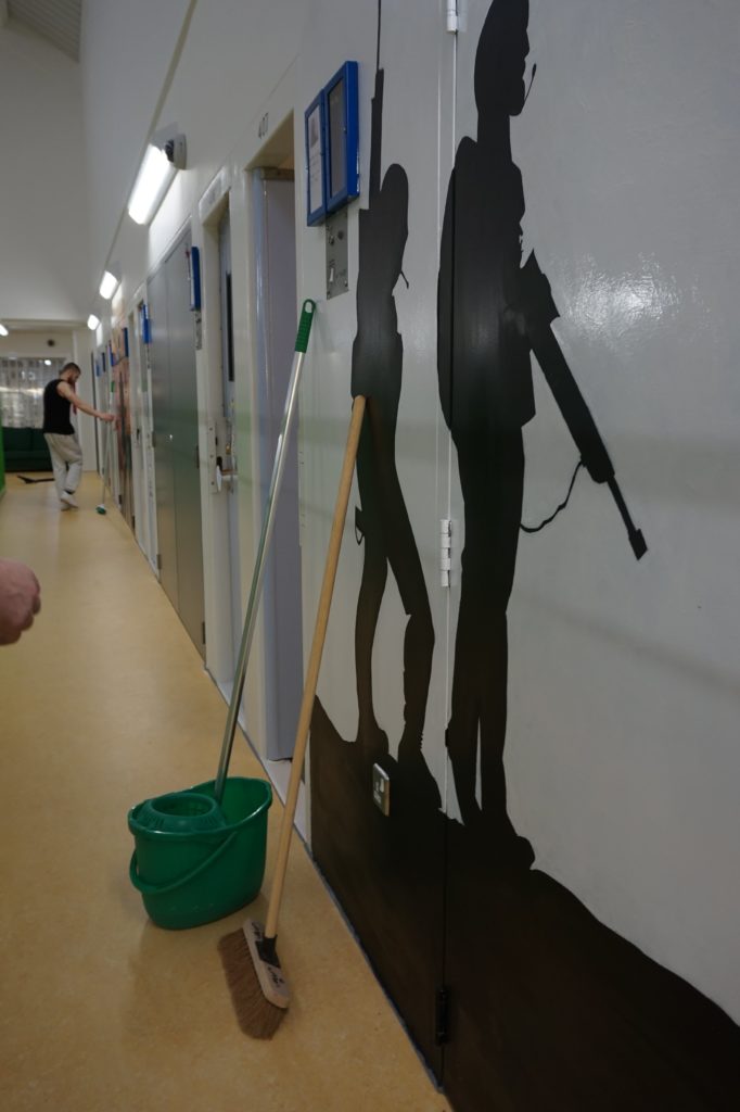 A mop and bucket lean on a wall that is adorned with painted silhouettes of soldiers, holding rifles, in a prison hall. To the left of the image are clasped hands of a prisoner staring at the soldiers, and in the background is a white male prisoner checking his prison money through a kiosk.