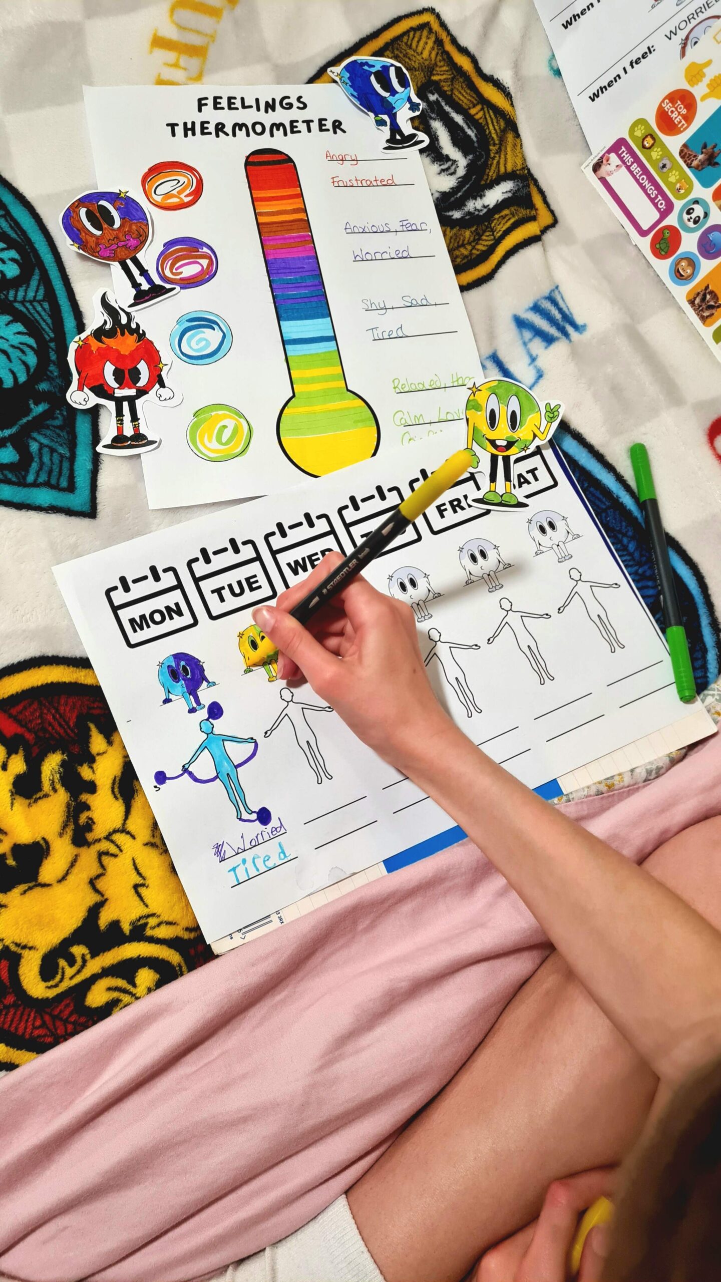 The image is a photo taken of a child completing two exercises from the PLUTO program: a psychological support program designed for children with Developmental Language Disorder. The top sheet is a Feelings Thermometer; the bottom sheet is a Weekly Emotion Tracker. The child has coloured in the feelings thermometer with the colours they associate with different emotions. For example, they associate yellow and green with happiness and light blue with tiredness. The child has also coloured in the cut-out PLUTO characters to match them with each emotion. The child’s hand is shown completing the ‘Tuesday’ on the Weekly Emotion Tracker, by first colouring in the PLUTO character, before drawing where the child feels the emotion in their body.