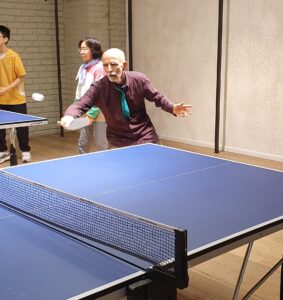 Older Caucasian gentleman playing table tennis in a community setting. Couple from Chinese community playing doubles table tennis in the background.