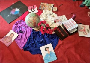 Teaching materials for a group workshop are laid out in a circle on a red carpet. Four coloured cloths are laid in a circle around a globe and a vase of red flowers to represent the four phases of the moon, which also relate to the phases of the menstrual cycle. On each of the four cloths there is a picture of a goddess and a coloured candle. Each phase is represented by a different colour cloth and the picture of a goddess. The new moon represents the menstrual period or red moon. The crescent moon is white and represents the follicular phase. The full moon is blue, full of energy and represents the ovulation phase. Finally, the waning moon is purple and represents the luteal phase. To the side of the picture, we can also see a picture of a uterus and a diagram of the moon phases. There is also a box of pamphlets for participants to take away and in the top right you can just see the feet of two women who participated in the group workshop.