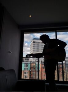 A silhouette of an asylum seeker playing his guitar inside a dark hotel room, with the colourful outside view behind.