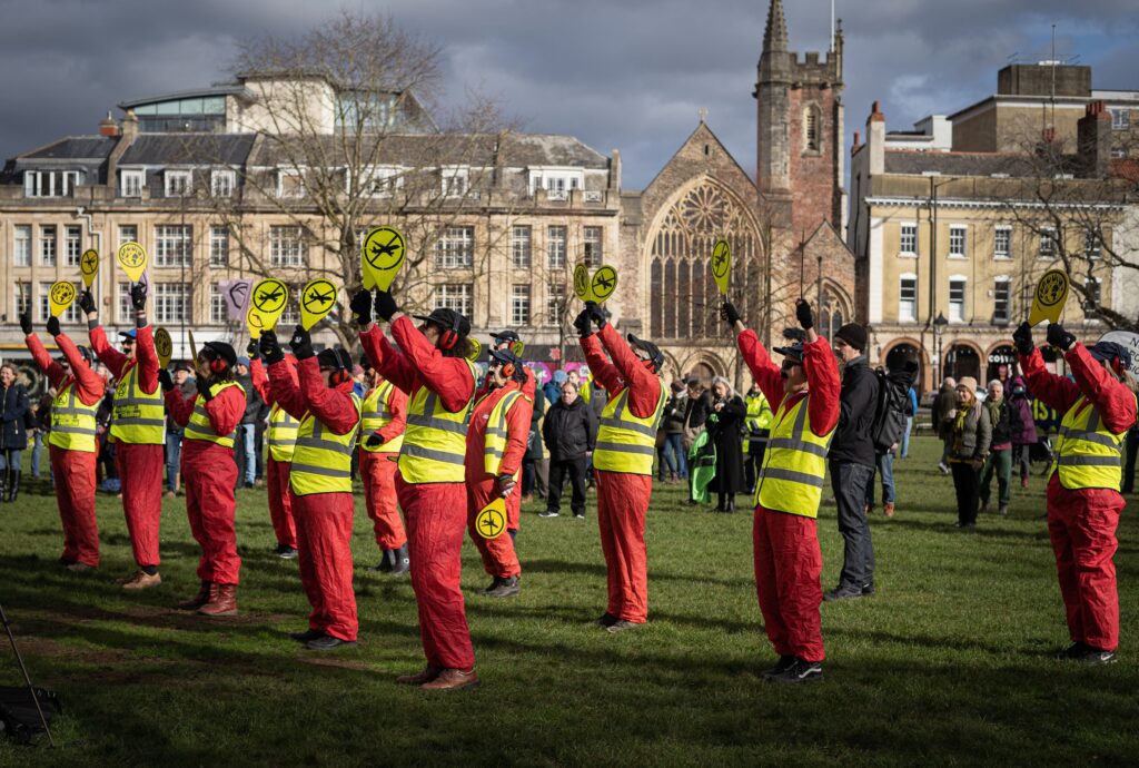 This image is of a group of 12 protesters dressed as air traffic controllers in high-vis vests holding up paddles with a vector of an airplane crossed out, stand on College Green, campaigning against the expansion of Bristol Airport. Several members of the public stand behind them looking on to the protest. 