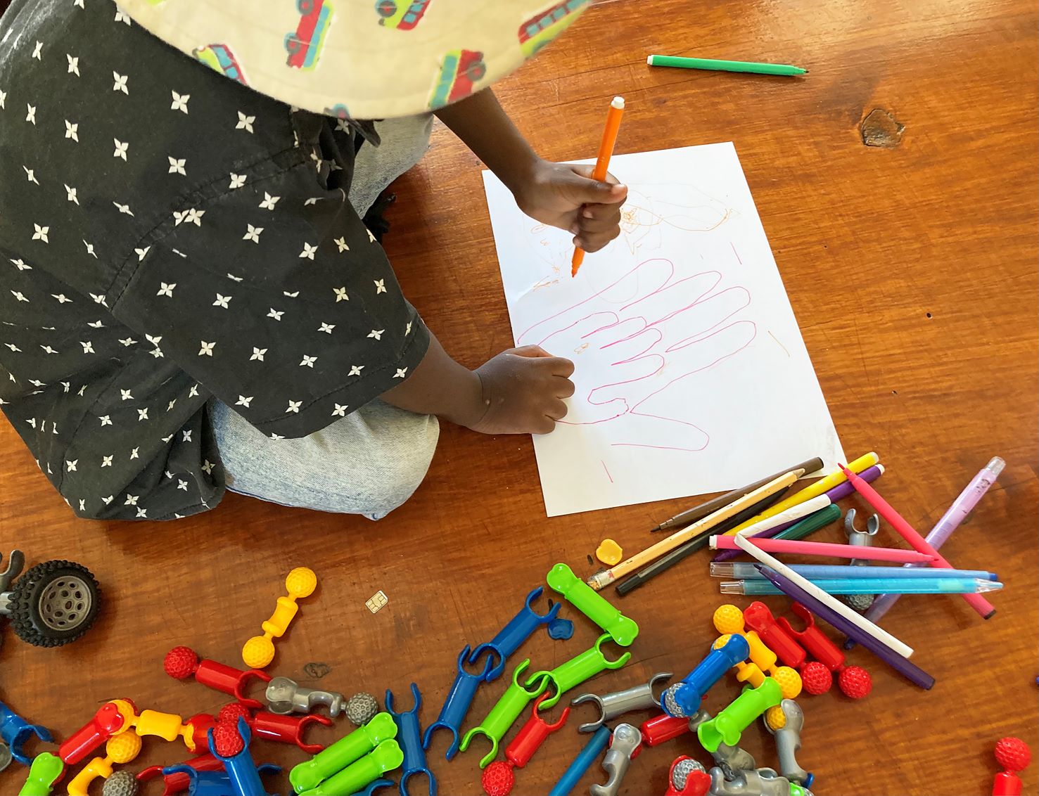 A young child sitting on a wooden table, drawing outlines of their hands on a piece of white paper with coloured pencils. Nearby, colourful building toys and pencils are scattered on the table. 