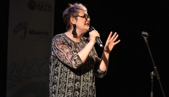A woman holding a microphone gesturing to the audience in front of a back background. There is a banner for the 'Minerva Lecture Series' behind her.
