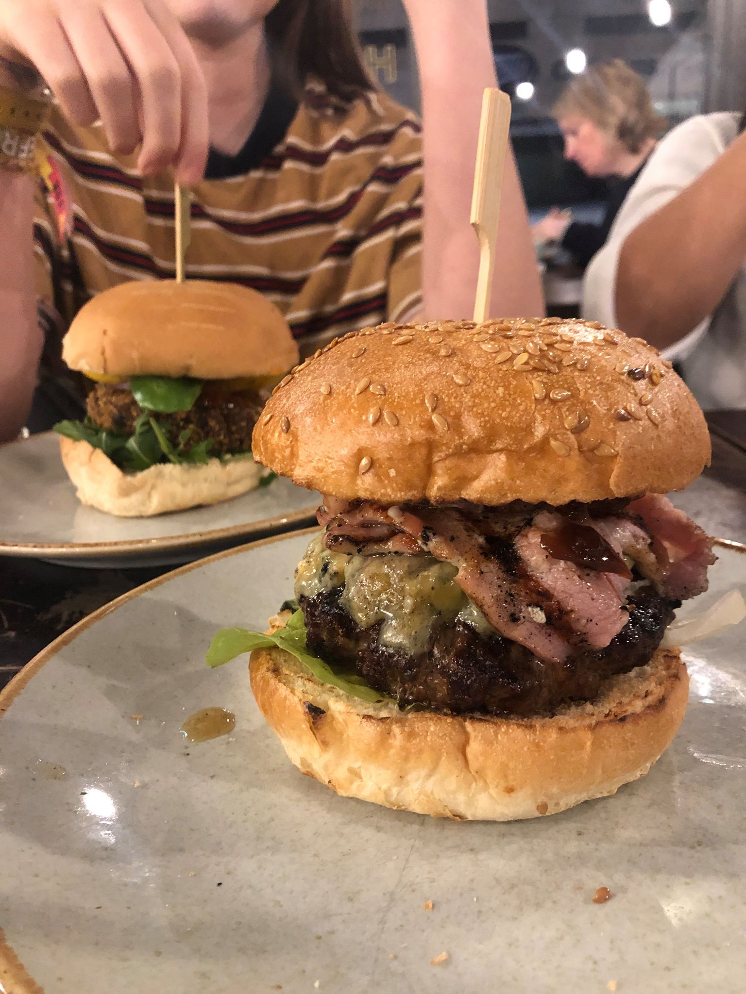 A lady sat in front of two bacon burgers on two separate plates in a restaurant. You can't see the person who will be eating the burger opposite hers.