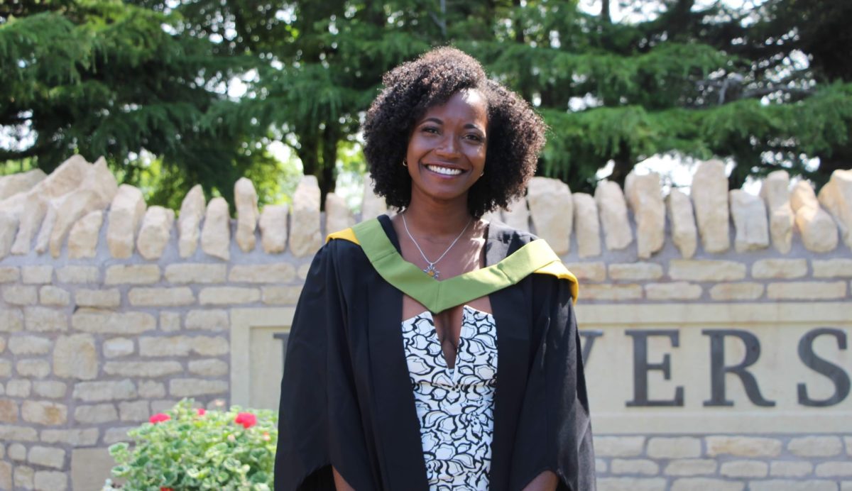 Toni wearing her gown stood inf front of the stone wall entrance of the University