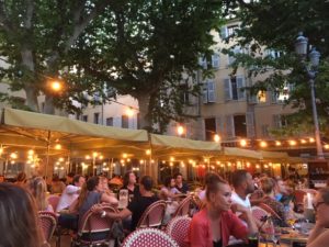 Pretty outside terrace in a French square, filled with parasols and fairy lights and lots of people eating and drinking at restuarant tables.