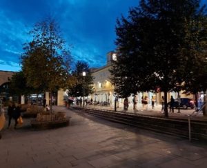 Bath City Centre with some green bushy trees inf the foreground and the buildings in the background. Some shoppers are on left-hand side of the photo