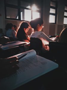 students sat in a hall undertaking an exam. The room is dimly lit by the sunset.