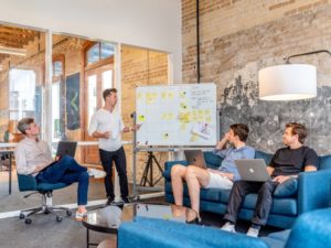 A group of 4 people sat on a sofa with a laptop and a big whiteboard filled with post-it notes. They are presenting to others not seen in the shot. 