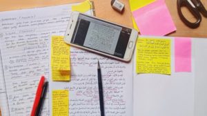 A mess of work placed on a table. Includes scribbled on yellow post-it notes, a phone with work on it, a pen and various books sprawled out. 