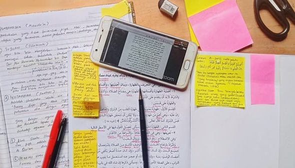 A mess of work placed on a table. Includes scribbled on yellow post-it notes, a phone with work on it, a pen and various books sprawled out.