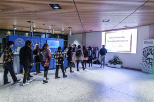 A group of students walking into the new School of Management building, in which projected on a wall is a powerpoint slide which reads 'Gold Scholarship Networking reception'